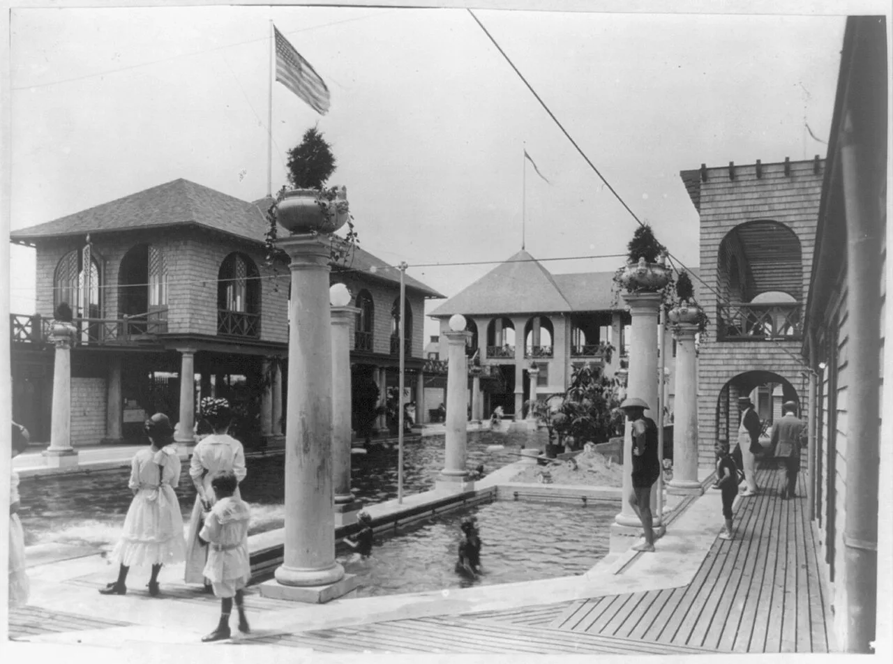 Early 1900s black and white photo of the pool court at the Monmouth Beach baths, with columns, a flag and visitors by the water