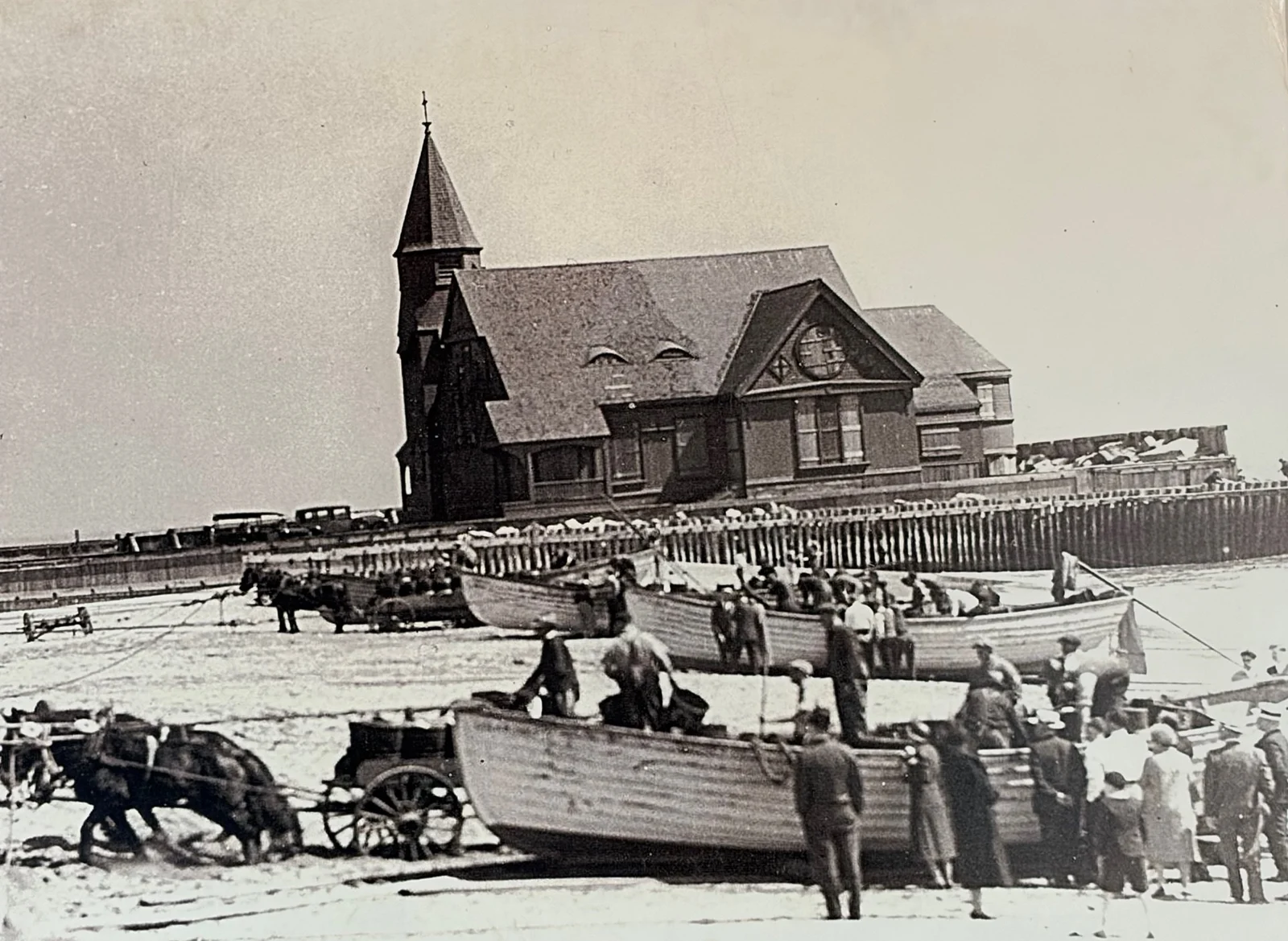 Black and white 1920s photo of wooden fishing boats hauled onto the beach by horses as onlookers gather, with St. Peter's Church of Galilee behind
