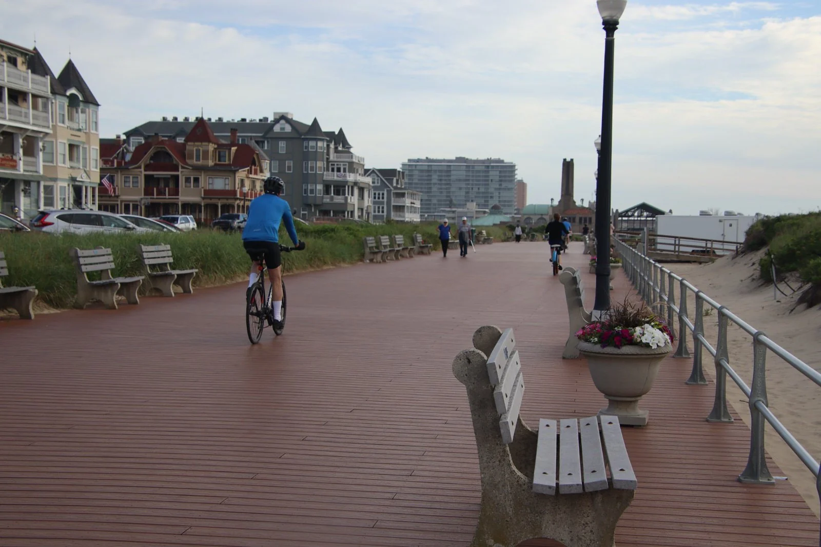 A cyclist riding along the Ocean Grove boardwalk past benches and flower planters, with Victorian buildings beside the dunes
