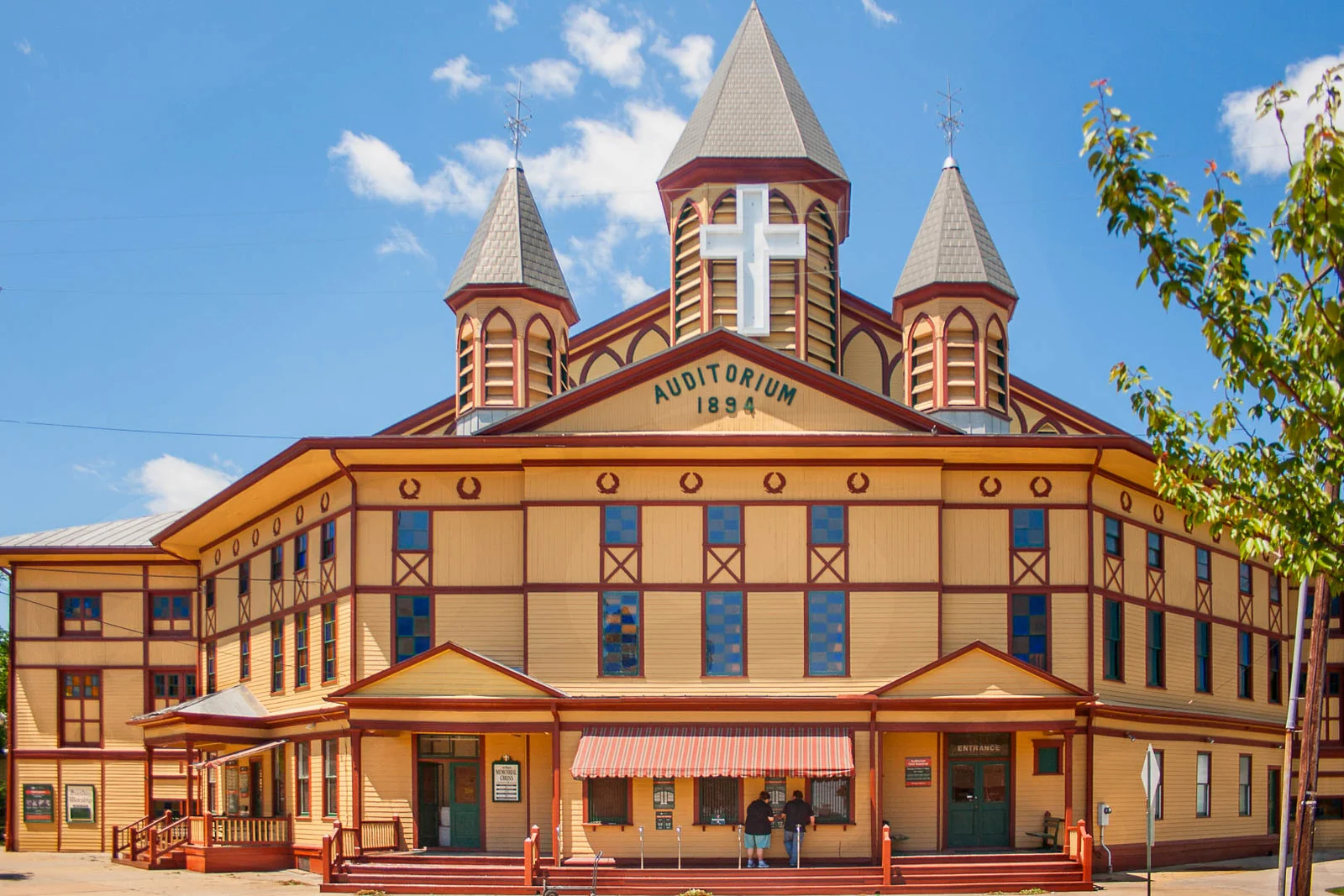 Front of the Great Auditorium, a yellow wooden hall with three spired towers, a cross and a sign reading Auditorium 1894