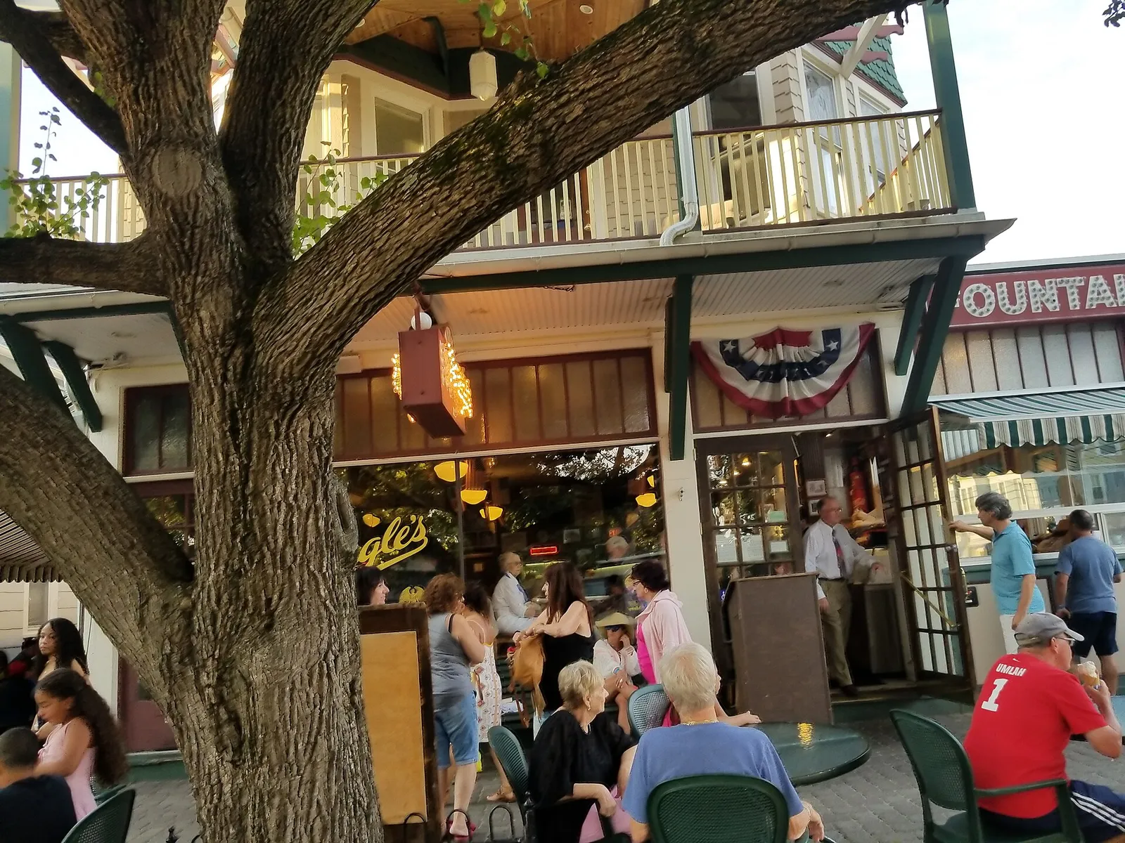 A busy Victorian storefront cafe with outdoor tables under a large tree