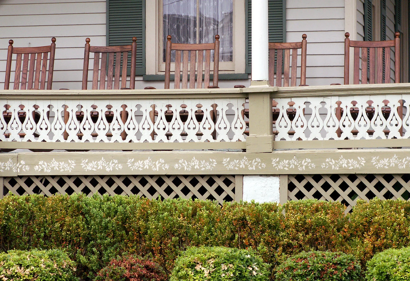 A row of wooden rocking chairs on a Victorian porch with a fretwork railing above green hedges