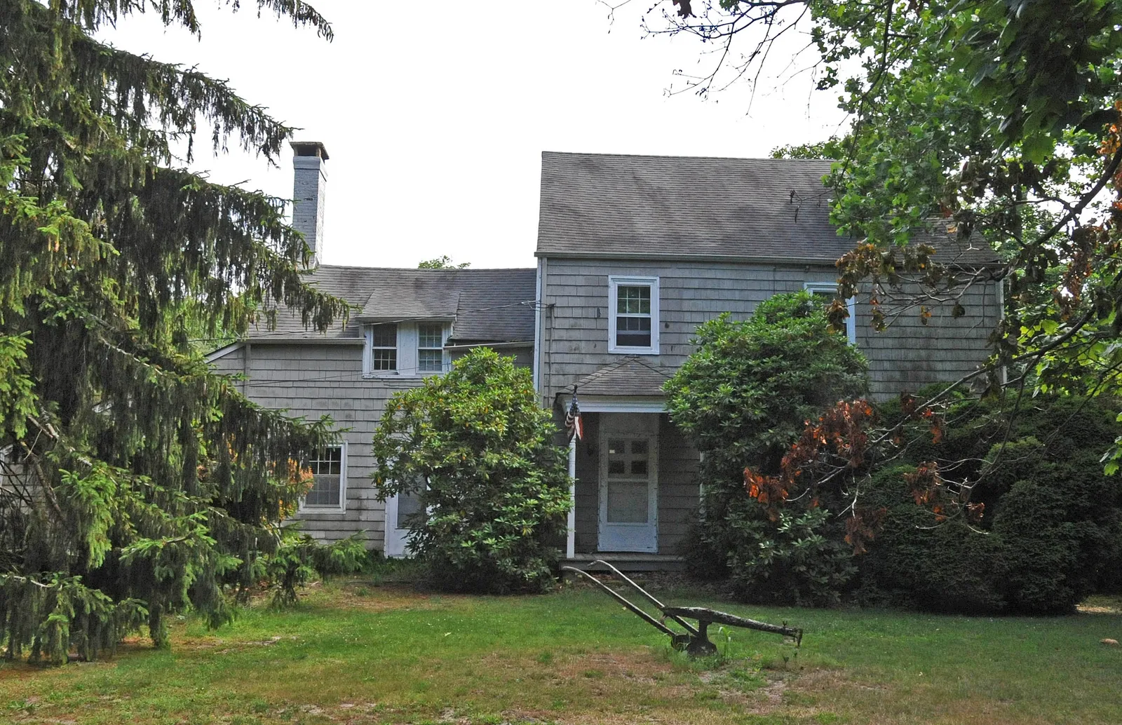 A gray shingled colonial farmhouse framed by evergreens, with an antique plow on the lawn