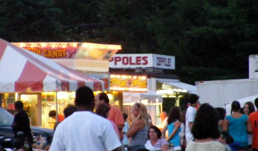 Evening crowd gathered by lit cotton candy and zeppole stands and a red and white tent at the township's July 4th celebration