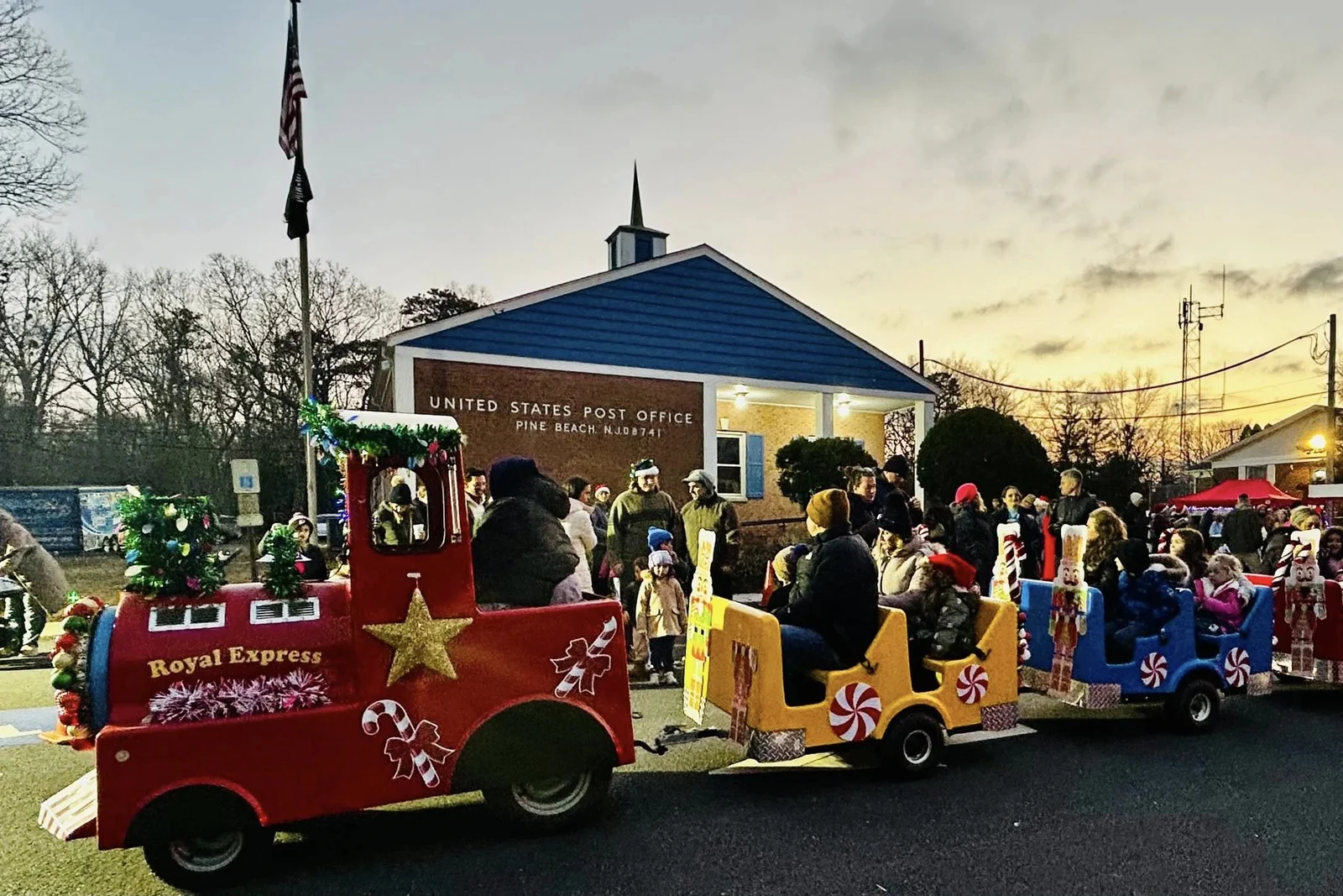 A decorated holiday train with a red Royal Express engine and candy-striped cars full of riders passing the Pine Beach post office at dusk