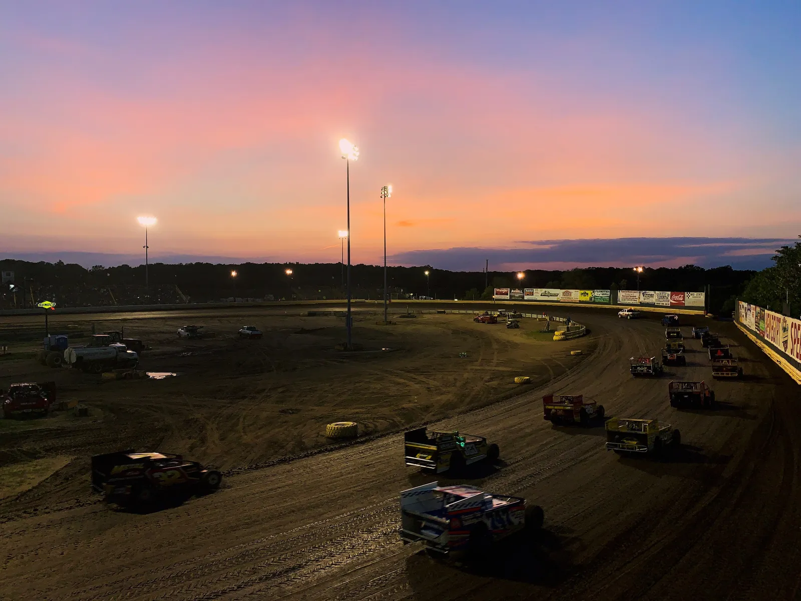 Dirt-track race cars rounding the oval under stadium lights and a pink sunset sky