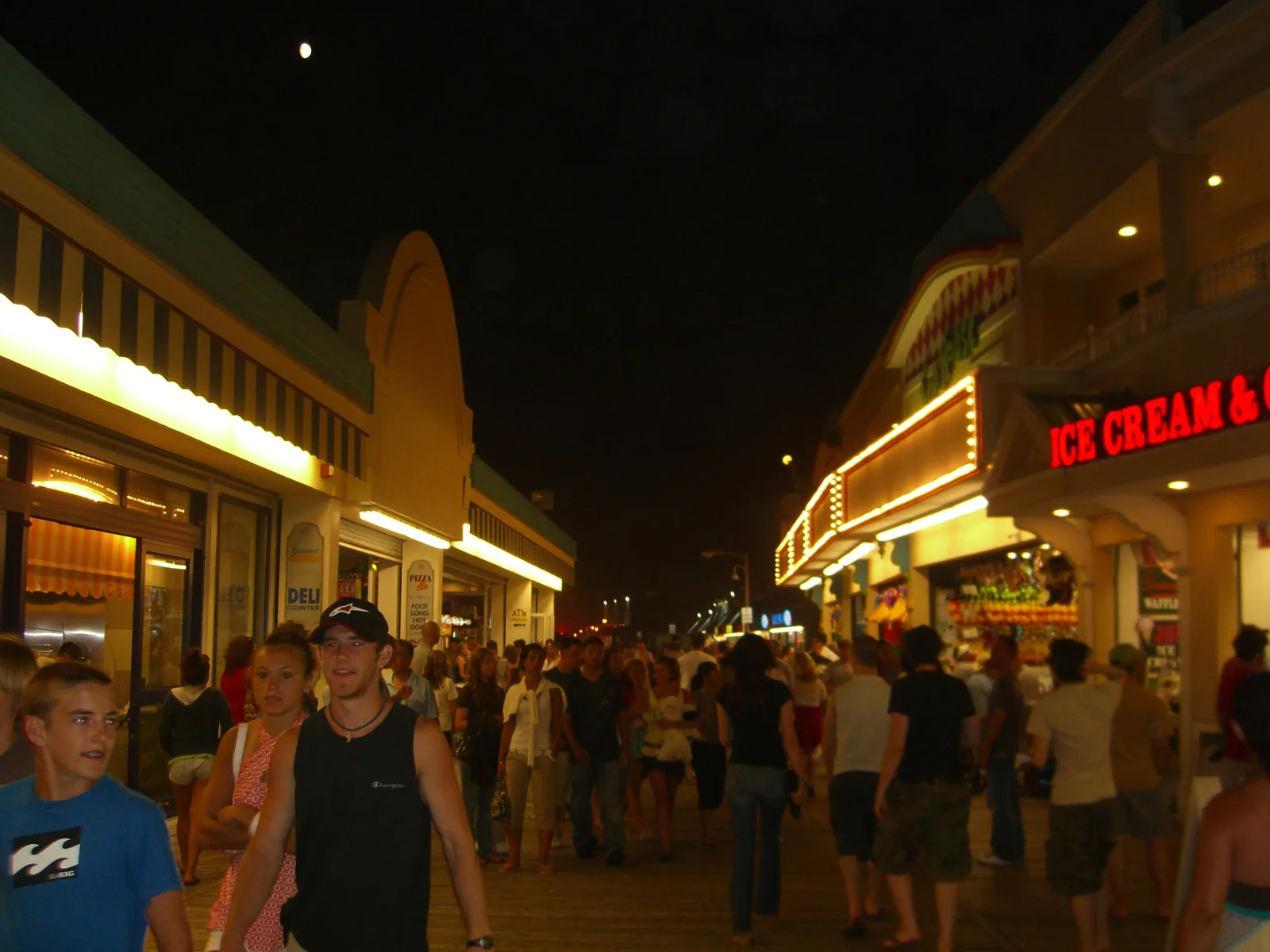 Crowds walking the lit-up Point Pleasant Beach boardwalk on a summer night, shops and ice cream stands on both sides