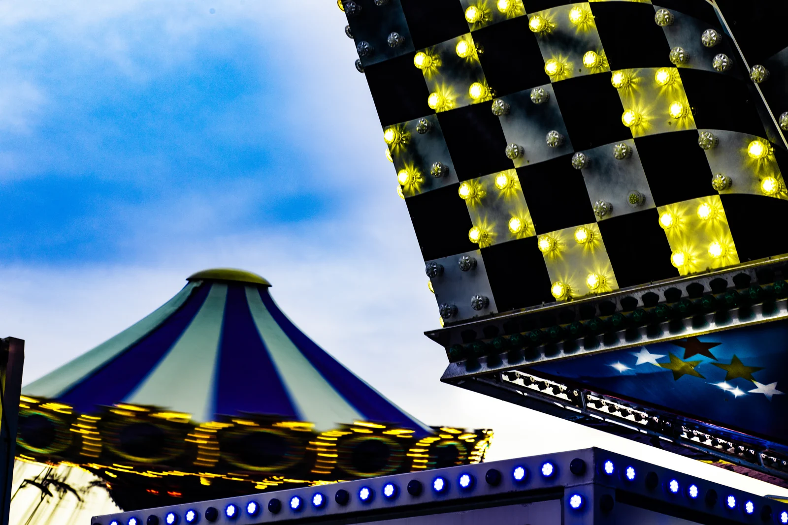 Checkered lights and a striped carousel canopy on boardwalk rides against a blue sky