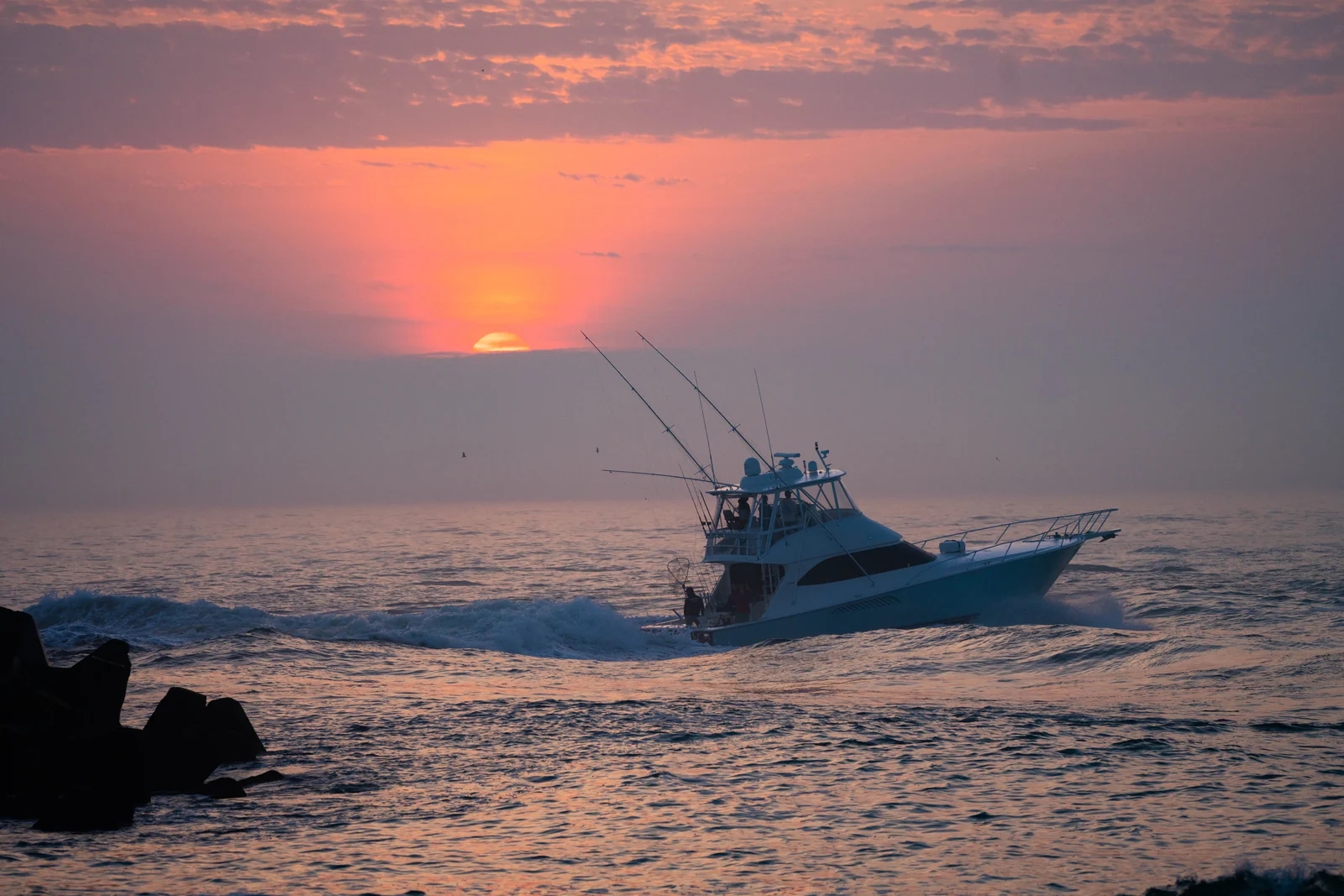A sportfishing boat with tall outriggers heading out of the Manasquan Inlet past the rock jetty at sunrise