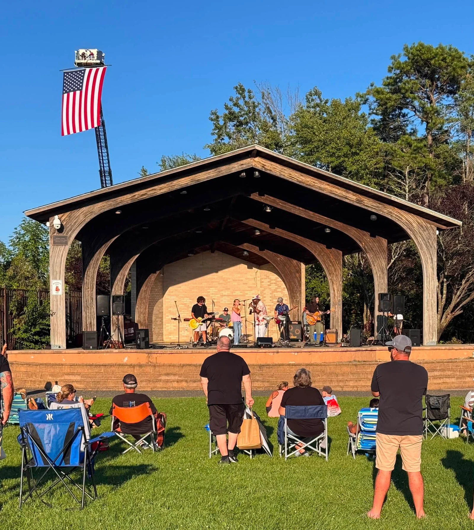 A band playing under a large open-air pavilion with an American flag flying above, while people in lawn chairs watch on the grass