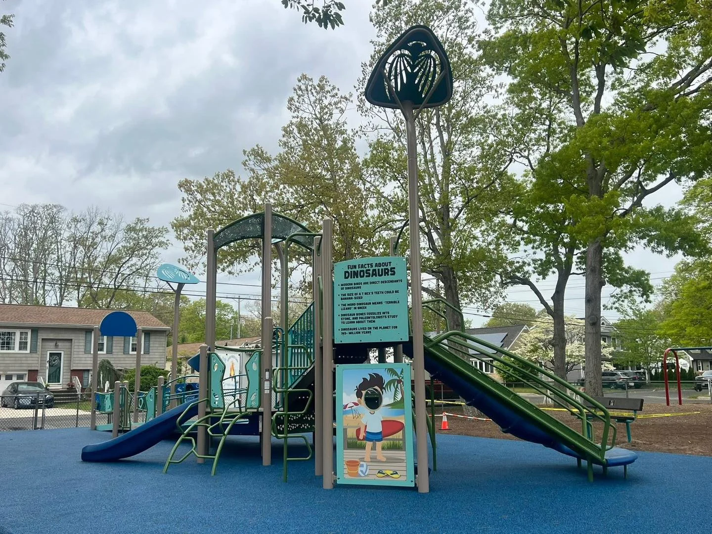 A new blue and green playground with slides, climbing towers and a shade canopy on a soft blue surface, surrounded by trees