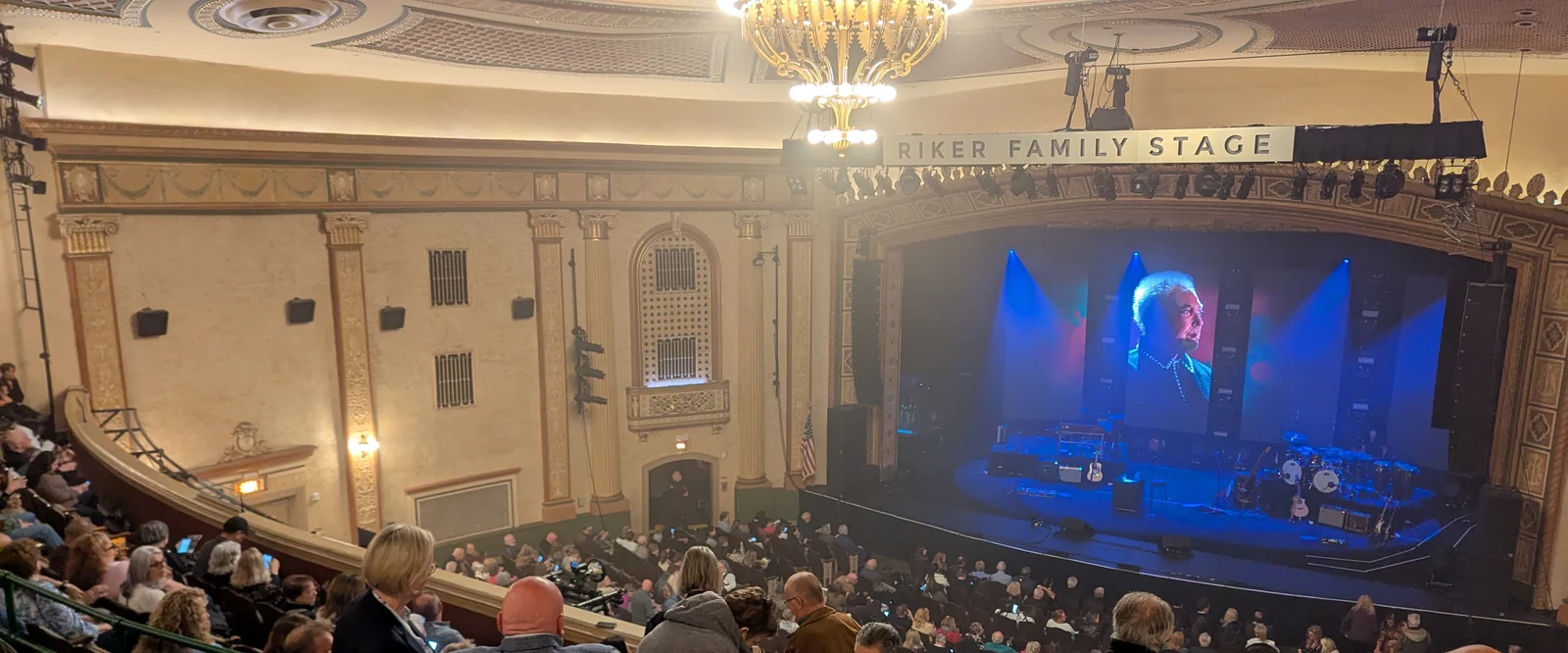 A packed Count Basie Theatre auditorium with gilded walls, a chandelier and a lit stage under the Riker Family Stage sign