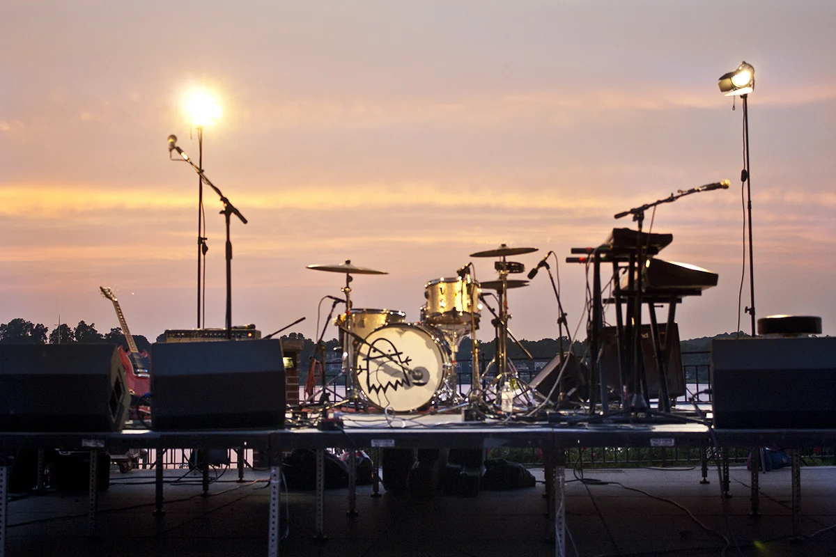 A band's drums, keyboards and guitars set up on an outdoor stage at sunset by the river