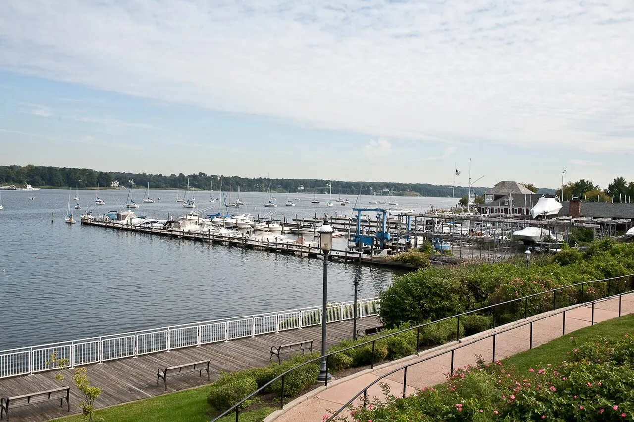 A brick riverside path with benches and railings overlooking docks full of boats on a wide calm river