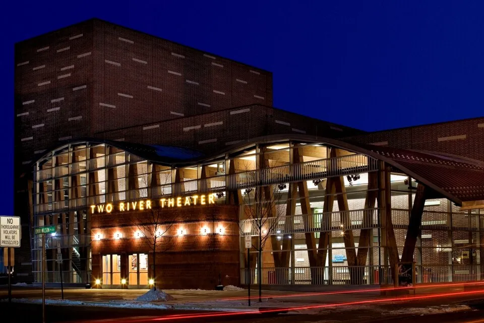 A modern brick theater with a curving glass and timber front lit up at dusk, its name in gold letters