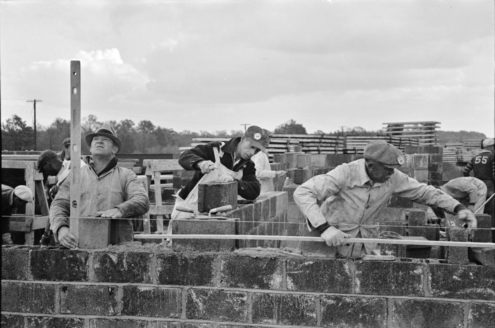Workers laying cinder blocks for a new homestead house at Jersey Homesteads, with stacked lumber behind them, November 1936