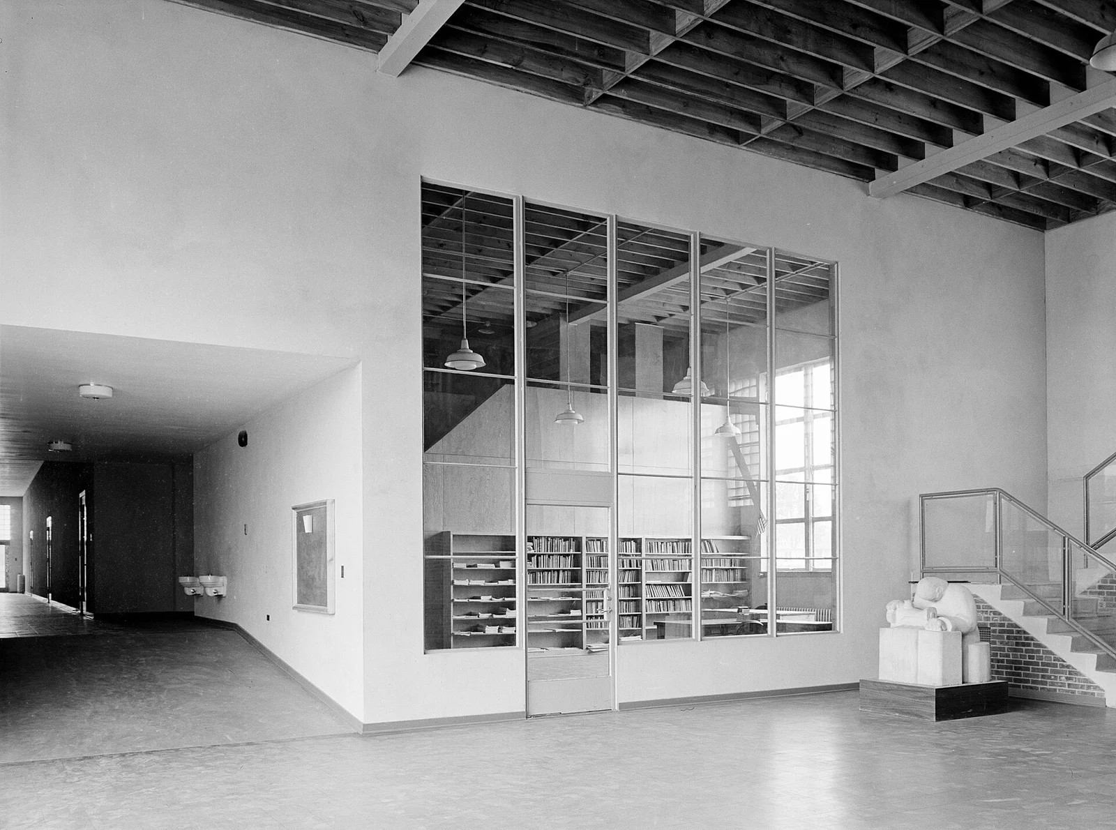 Bright modernist library room with a glass wall, bookshelves and a stairway beside a white stone sculpture, in the Jersey Homesteads community building, 1938