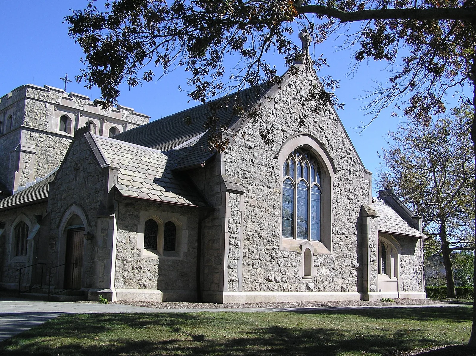 Gray stone Gothic-style St. George's-by-the-River Episcopal Church with a tall arched window, under shade trees