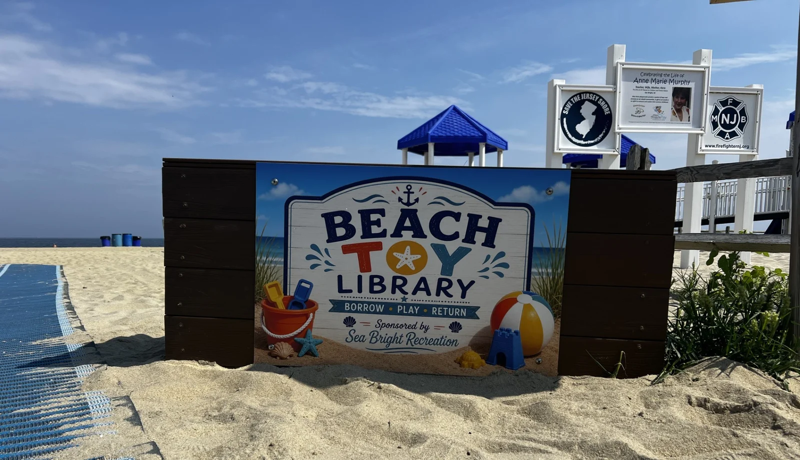 The Beach Toy Library sign on the sand at the Sea Bright municipal beach, beside a beach access mat and a blue lifeguard stand