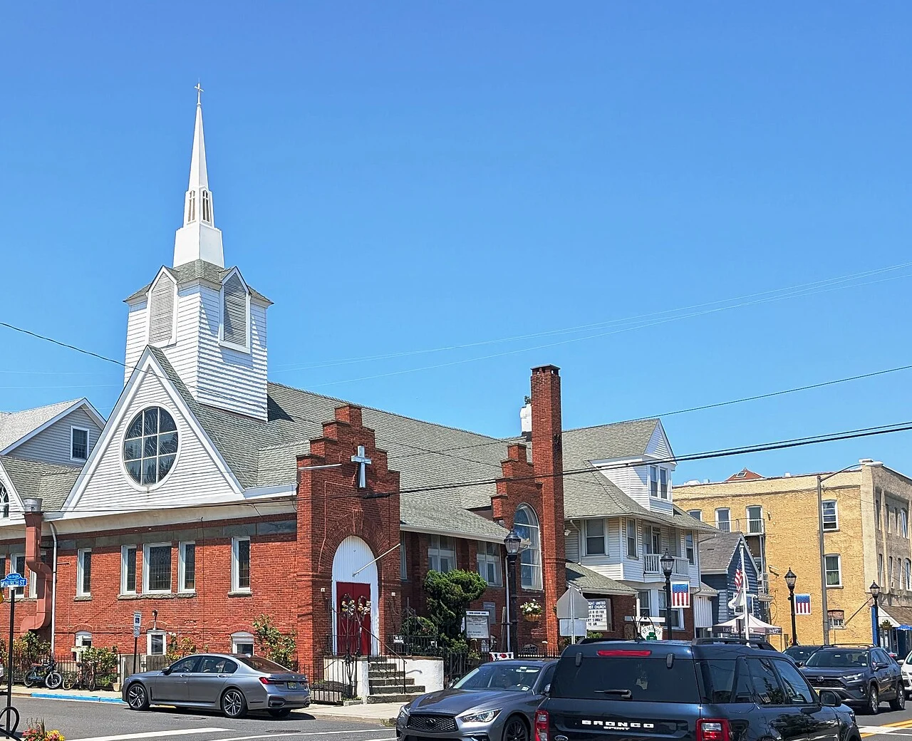 The brick and white clapboard Sea Bright United Methodist Church with a tall white steeple and a red door, on a sunny day