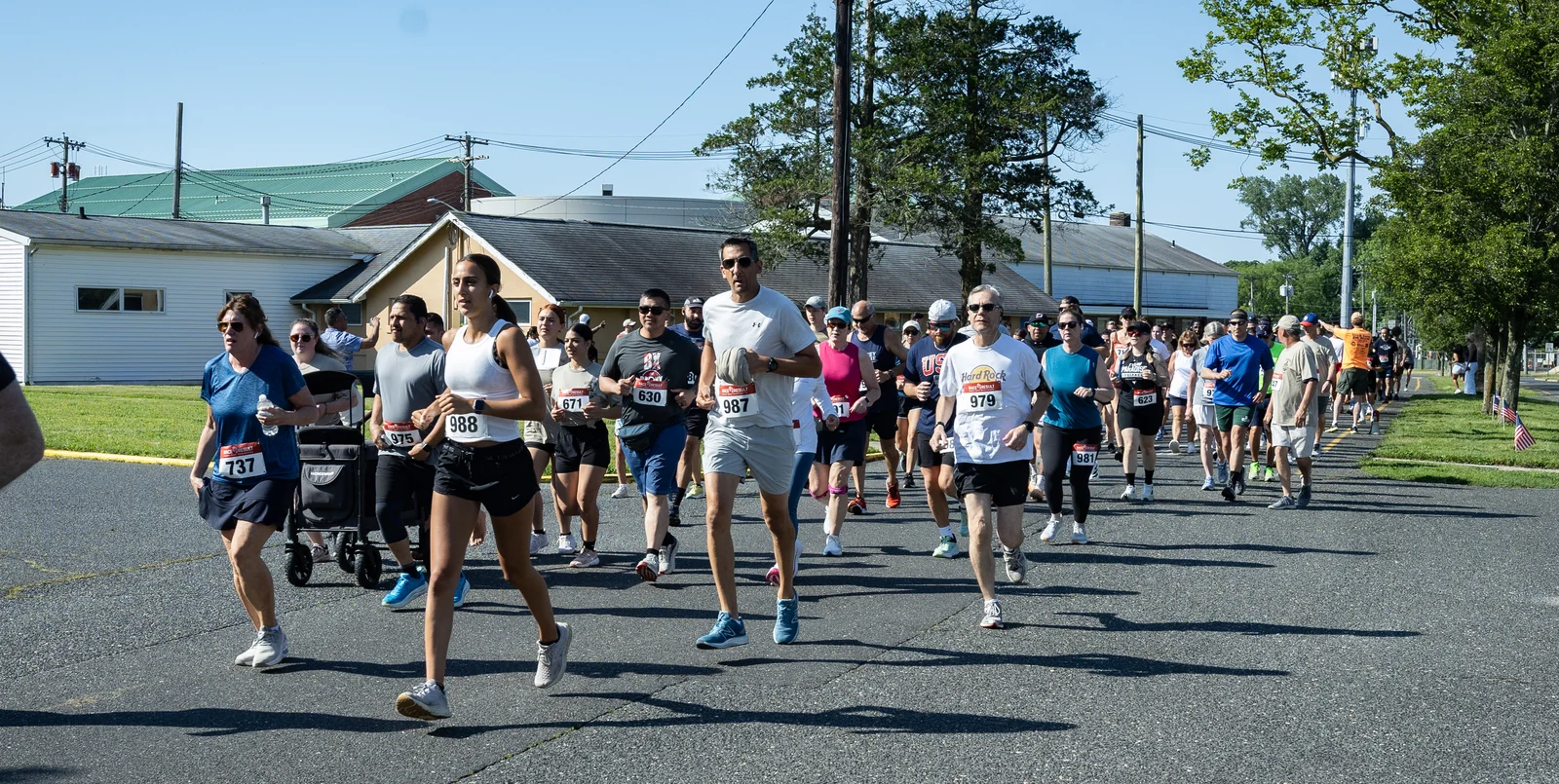 A large pack of runners in race bibs heading down a tree-lined road past low buildings on a sunny morning