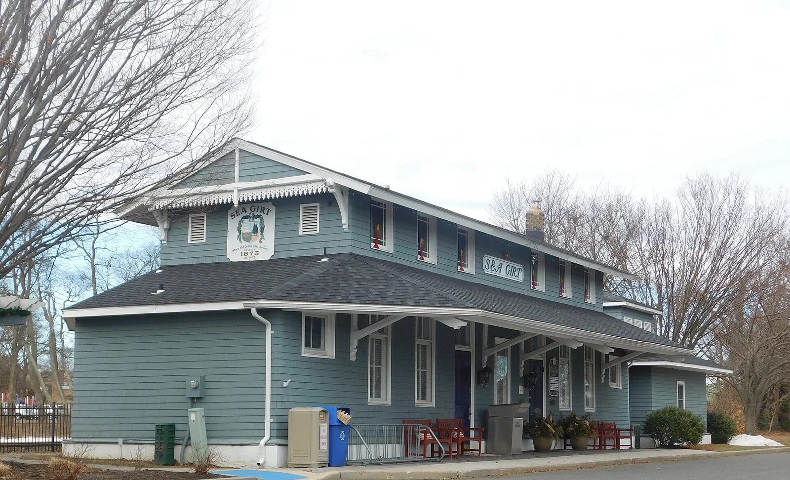 Blue-gray two-story former railroad station with a covered platform, red benches and a Sea Girt sign, now the borough library