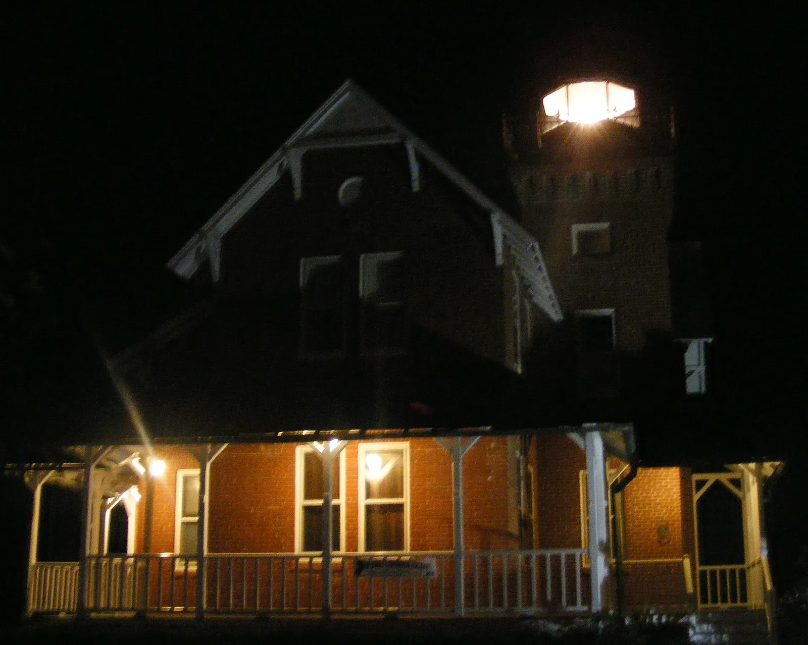 A brick Victorian lighthouse at night with its lantern glowing and the front porch lit warmly