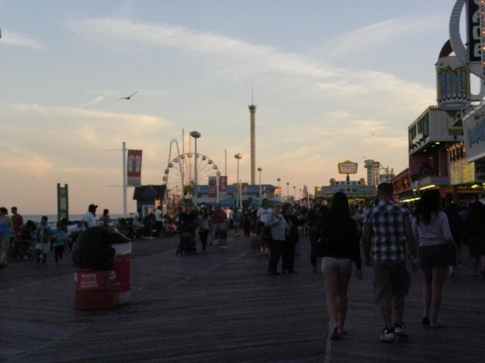 People walking the Seaside Heights boardwalk at dusk toward the Casino Pier Ferris wheel and rides