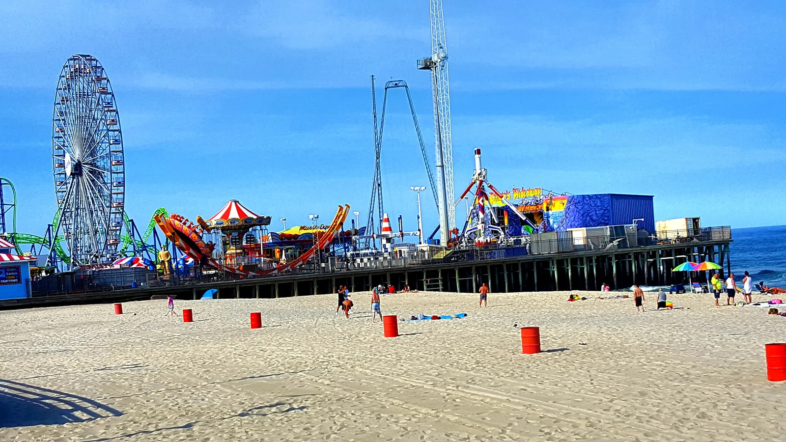 Casino Pier rides, including a Ferris wheel and the Hydrus roller coaster, seen across the sand under a blue sky