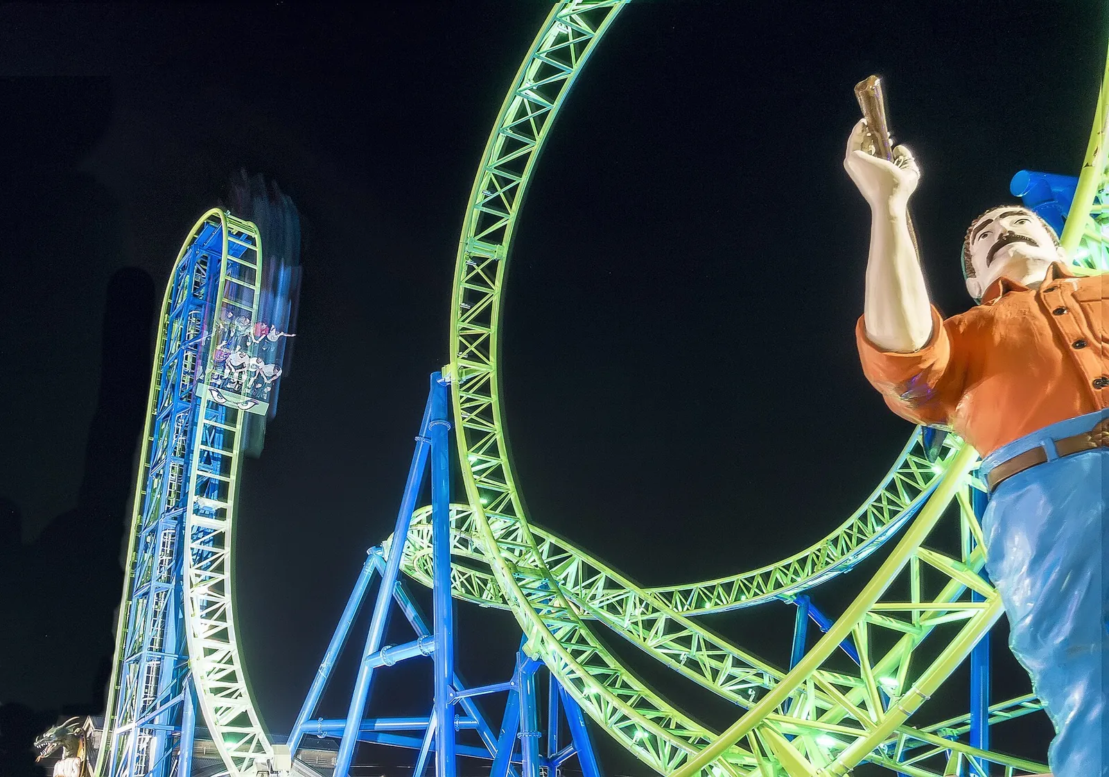The green and blue loops of the Hydrus roller coaster lit up at night beside a large statue on Casino Pier