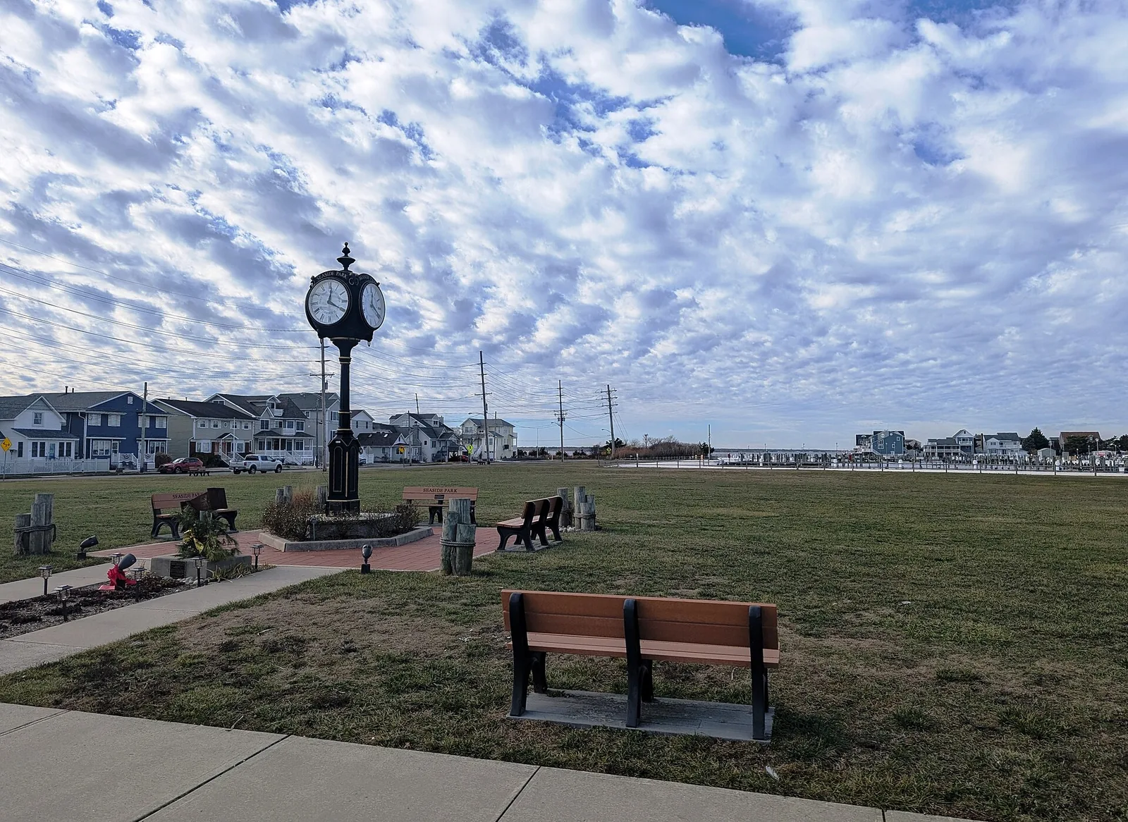 A tall street clock, benches and a green lawn at Berkeley Harbor Marina Park, with bayfront homes behind