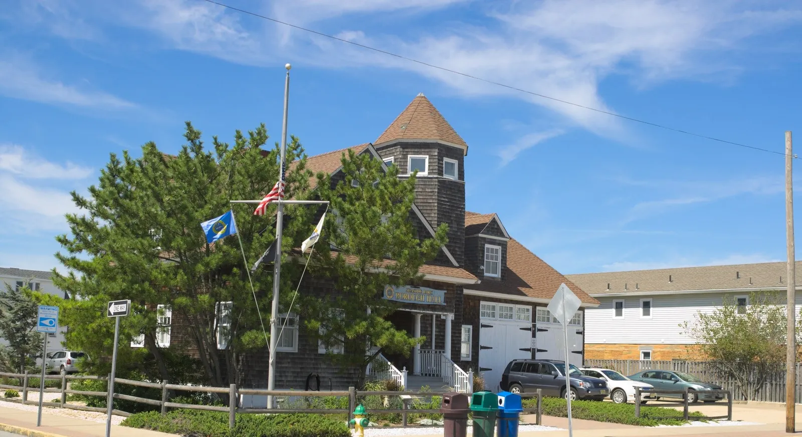 A brown-shingled seaside building with a pointed corner tower, flagpole and evergreen trees under a blue sky