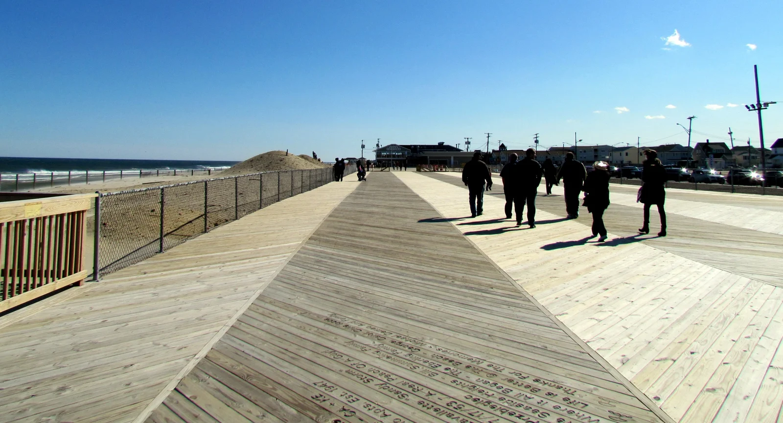 Wide wooden boardwalk in Seaside Park under a clear blue sky, with people strolling toward the Park Pavilion and the dunes on the left