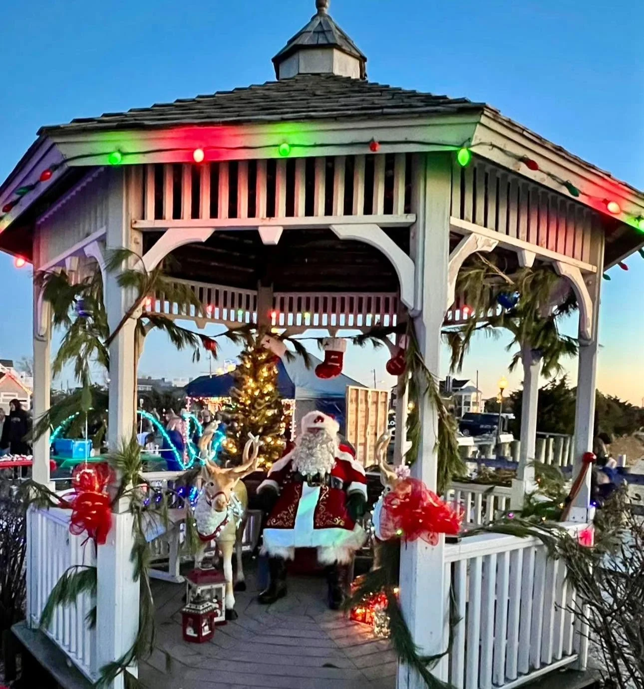 Santa seated in a white gazebo trimmed with red and green lights, garland and stockings, beside a lit Christmas tree and light-up reindeer at dusk