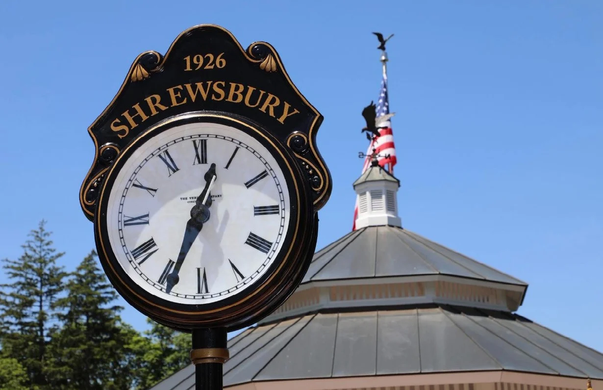 The new black and gold 1926 Shrewsbury street clock in front of the town gazebo roof and an American flag under a blue sky