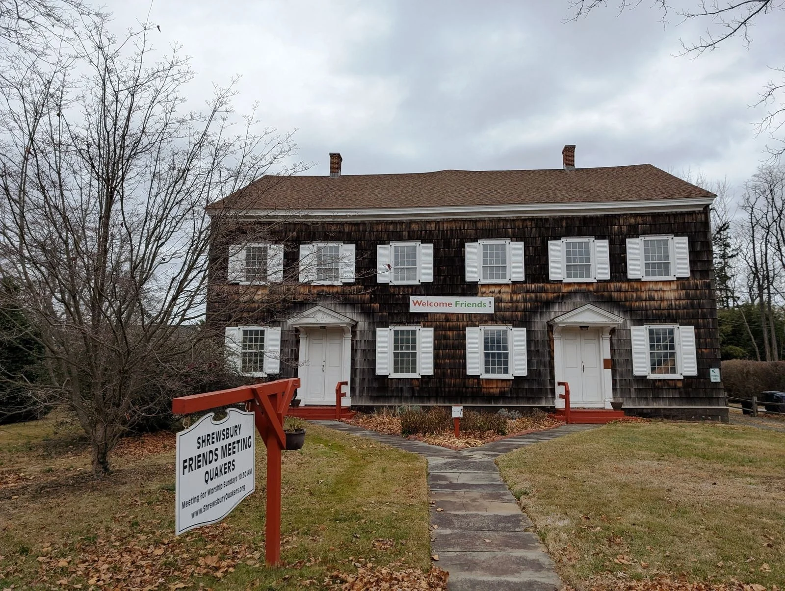 The dark-shingled Friends Meetinghouse with white shutters and twin doors behind its sign on Sycamore Avenue
