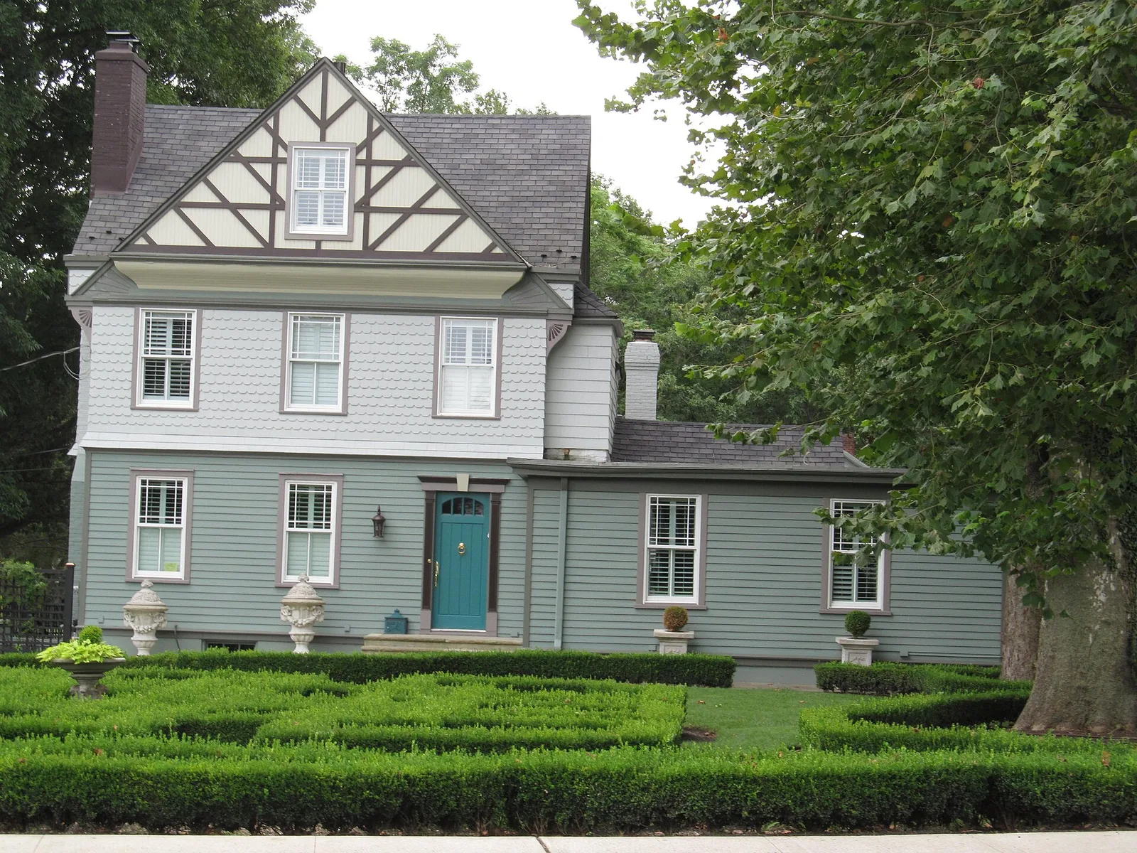 A gray home with a half-timbered gable and a teal door behind a clipped boxwood garden