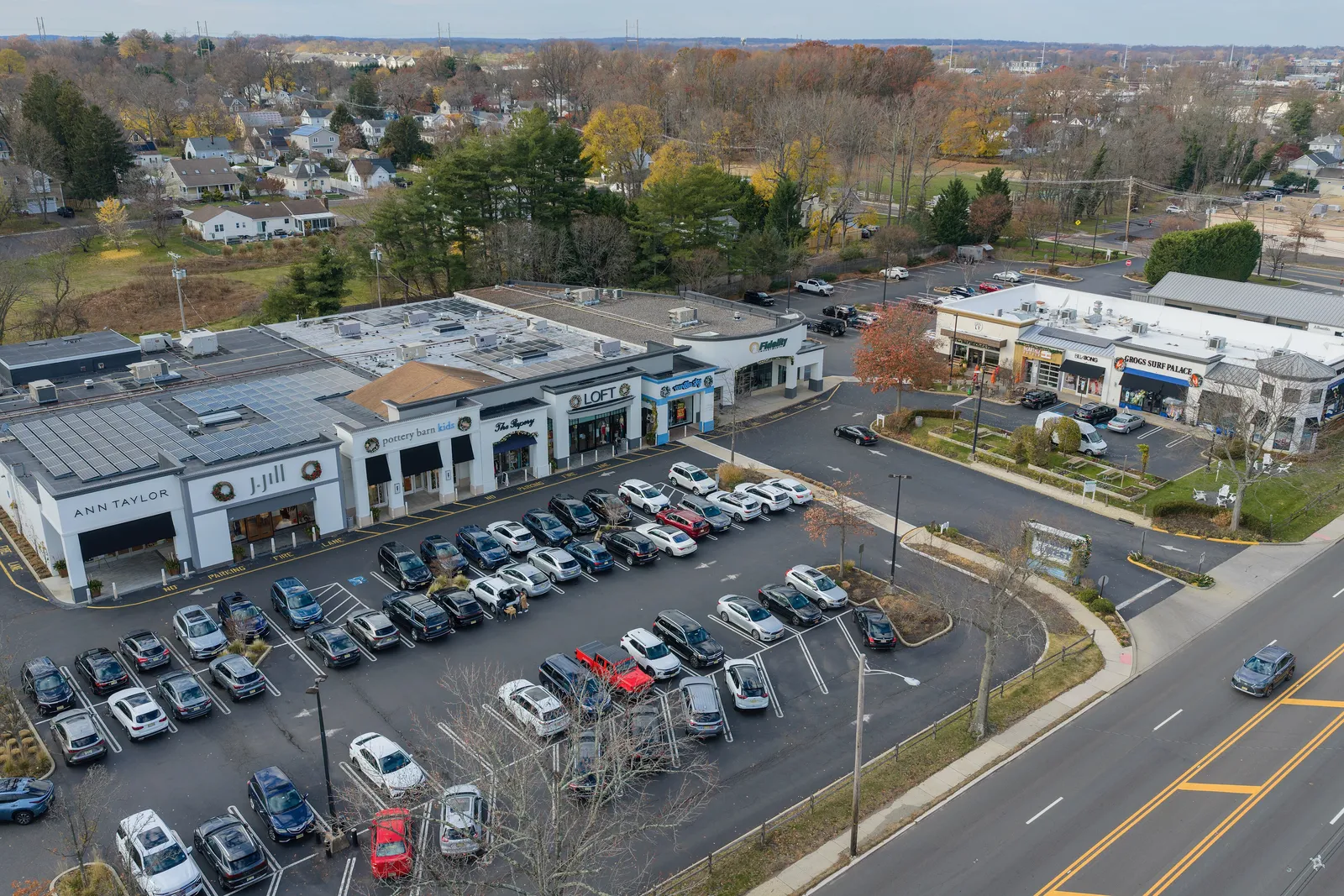 Aerial view of an open-air shopping center with shops and autumn trees beside a main road and neighborhoods beyond