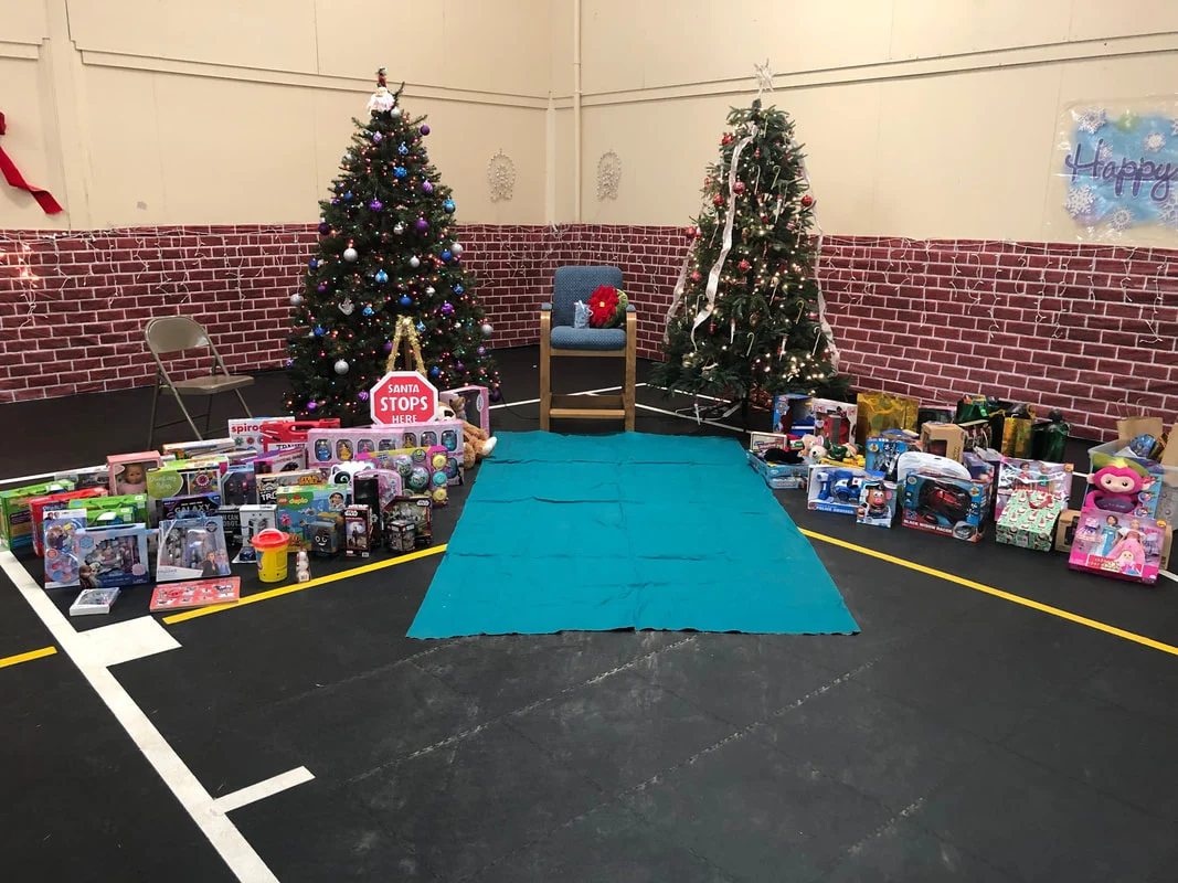 Township gym decorated for the holidays with two lit Christmas trees, a Santa Stops Here sign and rows of wrapped toys around a chair for Santa