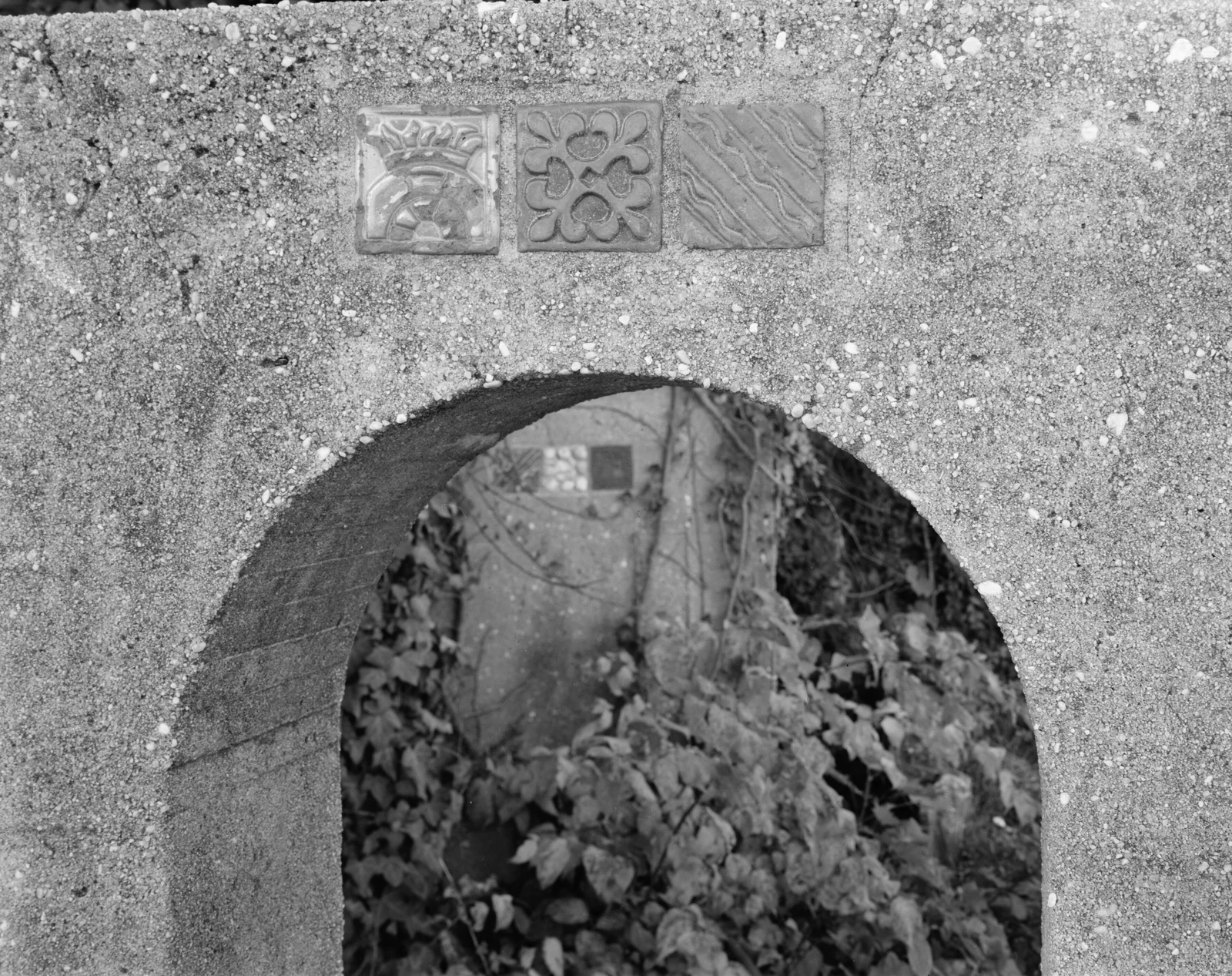 Black and white close-up of a cement arch with three decorative tiles set above it, one showing a crown, with ivy growing behind