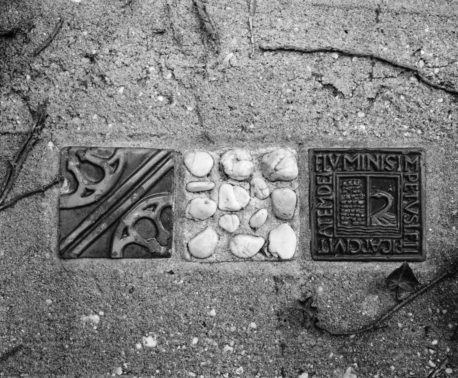 Black and white close-up of three decorative tiles set into the Birdville cement arcade, one with a Latin inscription
