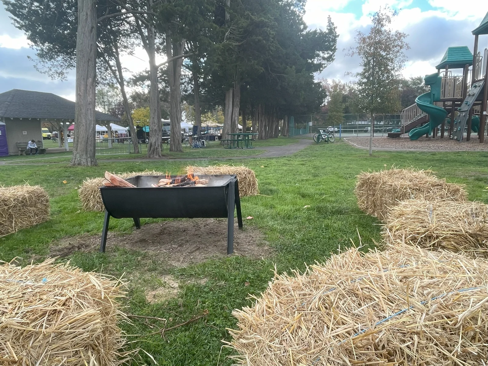A steel fire pit ringed by hay bales, with vendor tents, a pavilion and a playground beyond