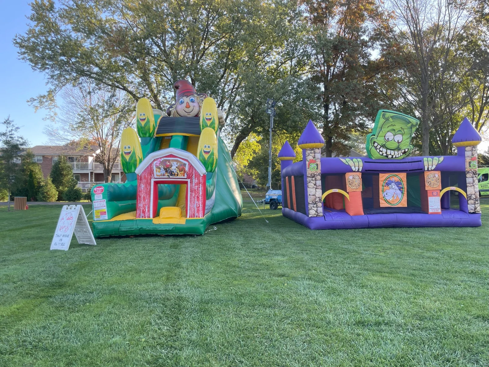A corn-themed barn inflatable slide and a purple castle bounce house on the grass under autumn trees