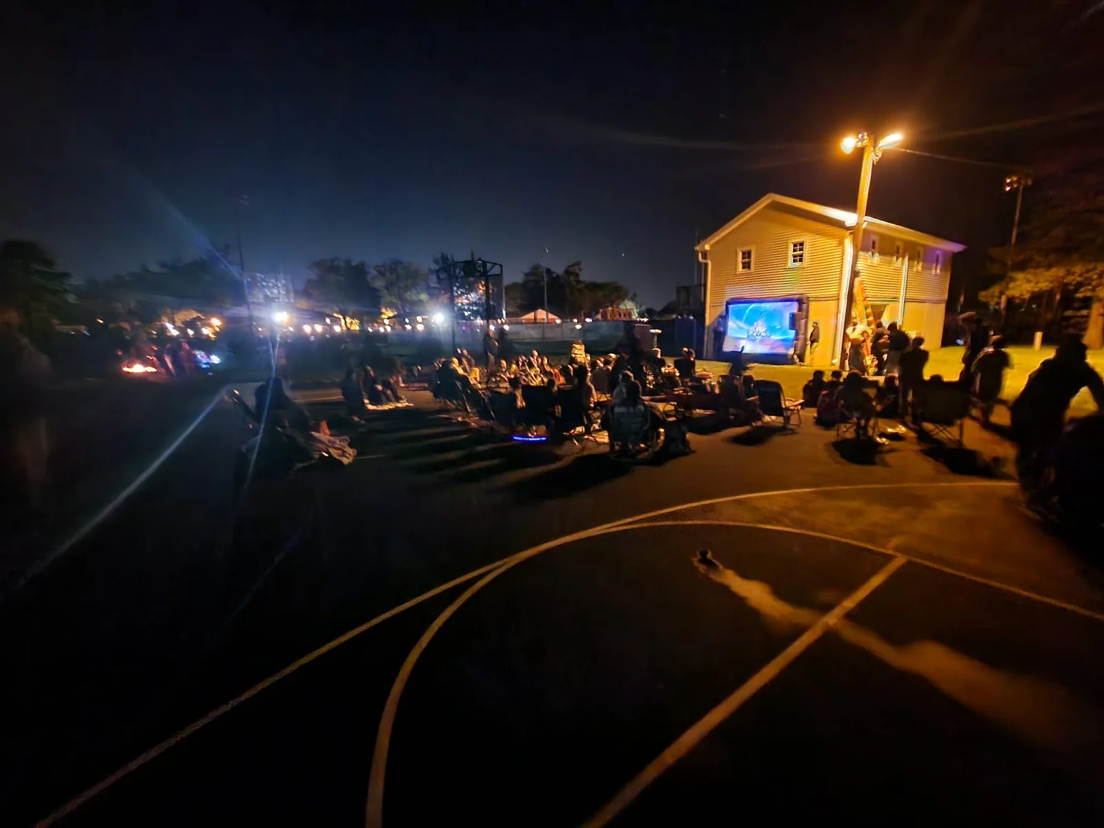 Crowd in lawn chairs watching a film on a large outdoor screen at night, beside a lit yellow building