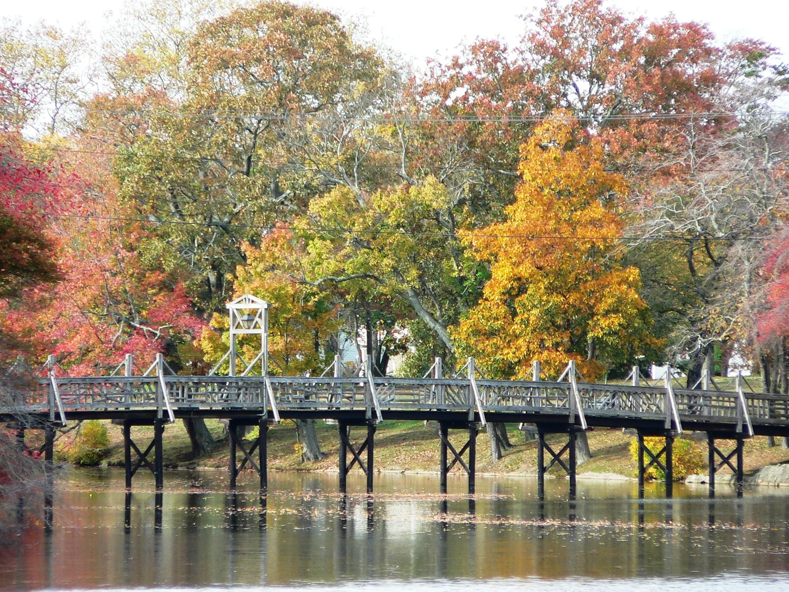 A white wooden footbridge crossing the lake beneath trees in red, orange and gold fall color