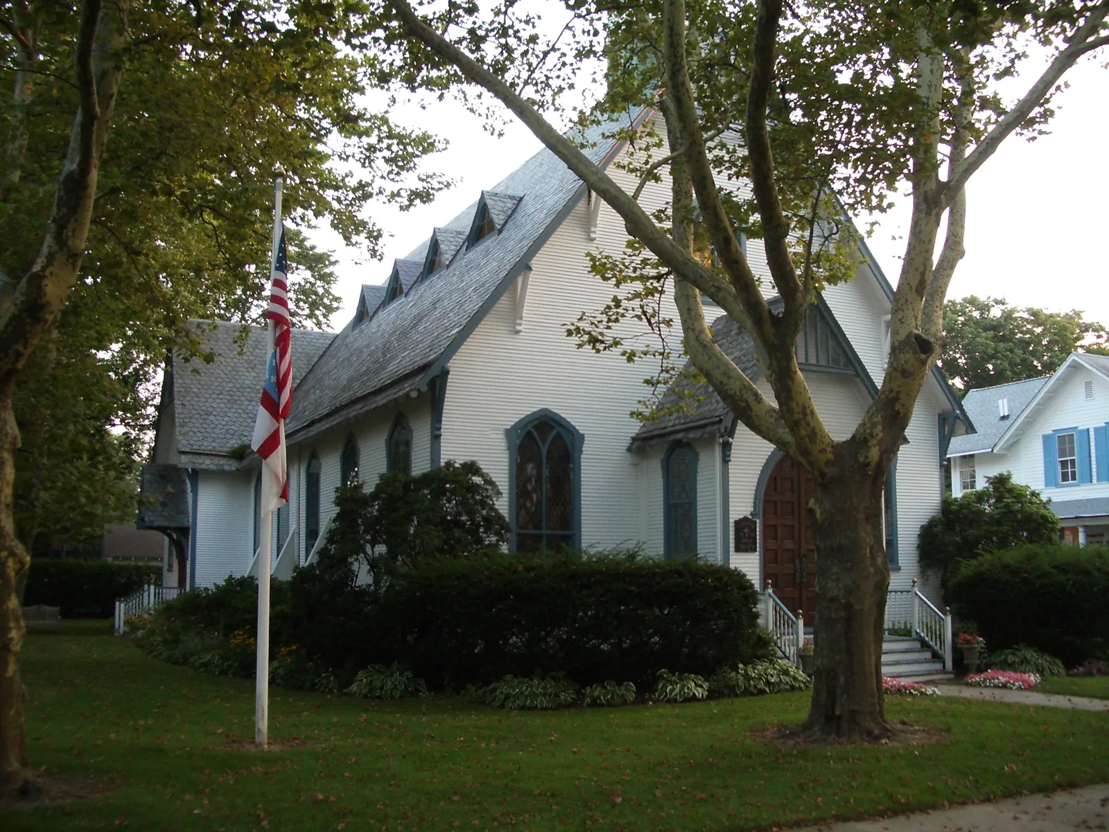 A white shingled Gothic Revival church with pointed windows, shaded by tall trees with a flag out front