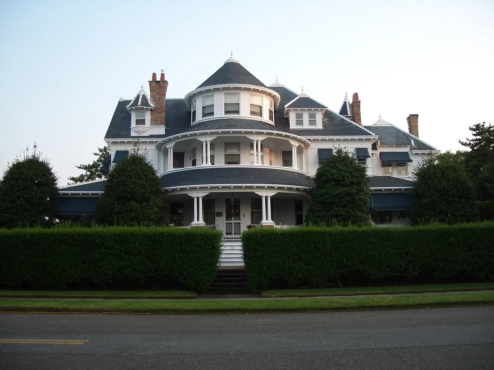 A large gray Victorian house with a wraparound porch, a round turret and dormers behind a trimmed hedge