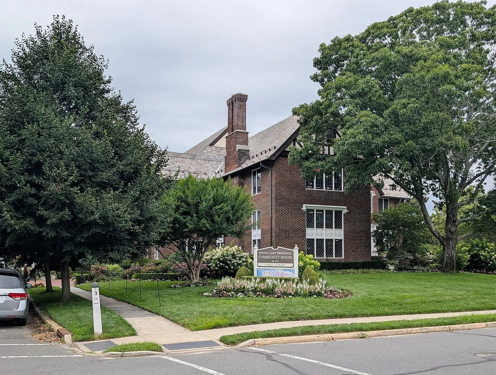 The brick Spring Lake Memorial Community House with its steep slate roof, framed by leafy trees and flower beds