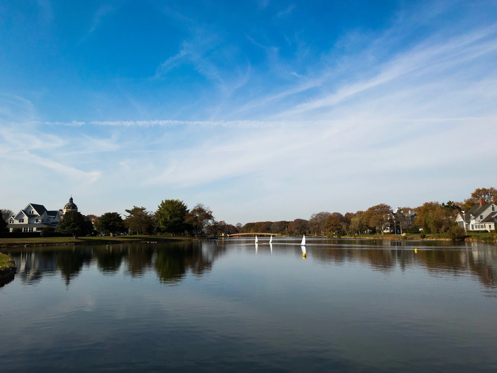 Small white model sailboats on the calm lake under a blue sky, with houses and trees reflected on the far shore
