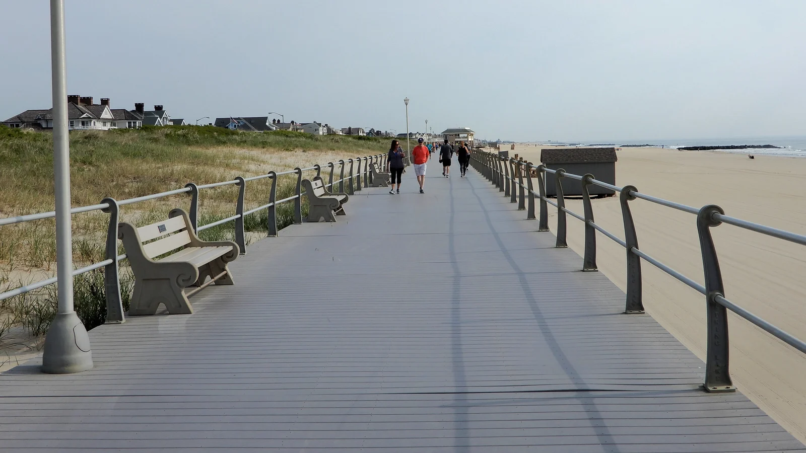 The wide wooden boardwalk with benches and railings running alongside the dunes and beach, with people walking in the distance